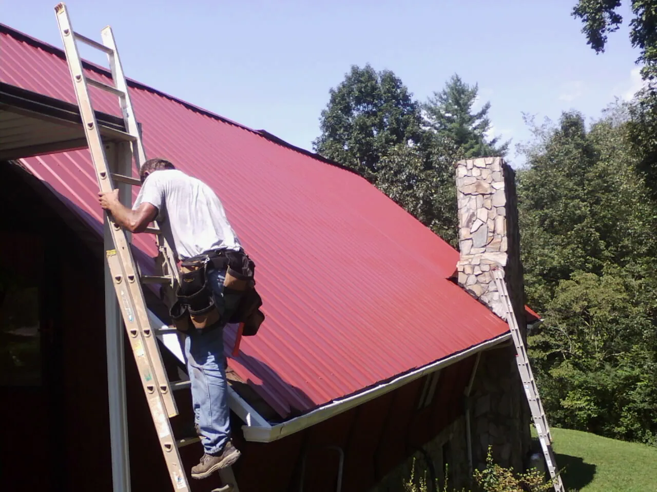 Roofer on a ladder beside a red metal roof and stone chimney — Soffit & Fascia Repair by Roofing Brothers in Dalzell